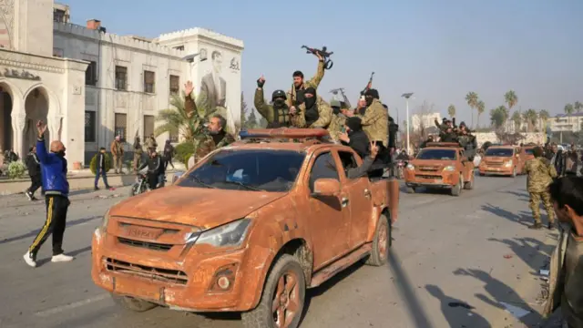 A group of male fighters sit on the back of a vehicle, with guns raised, as it passes through the street in a convey with others