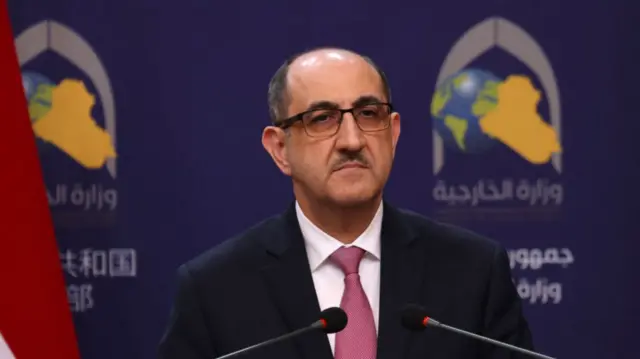 Hassam al-Sabbagh attends press conference with his Iraqi and Iranian counterpart. Mid-shot, cuts off below the shoulders: he's standing in front of a podium wearing a black suit and pink tie, behind him is a blue wall with a logo of the world, a grey canopy an stylised map of the middle east