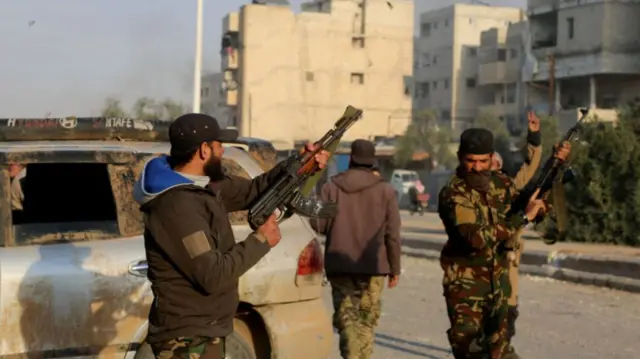 Two Syrian rebles stand in front of a destoryed car, pointing shotguns into the air.