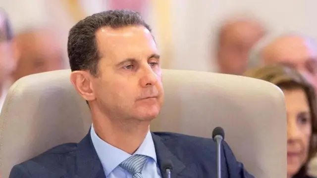 Close up shot of Bashar al-Assad wearing a blue suit. There is a small microphone in front of hi,