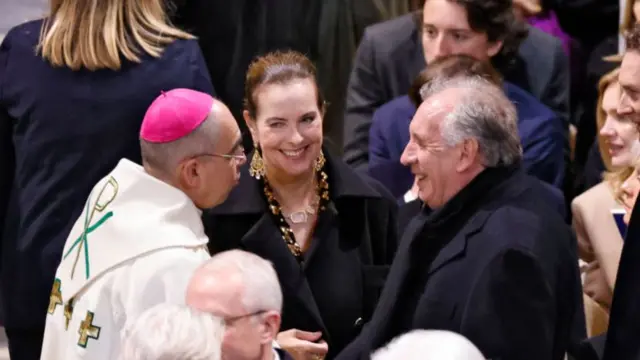 Carole Bouquet (C) smiles as she speaks to other guests. She has her hair slicked back and is wearing a black coat and heavy golden earrings with matching necklace