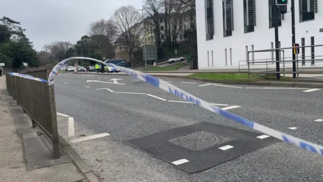 The A390 at the PKF Francis Clark building in Truro is empty except for a police car. There is police incident tape flapping between barriers in the foreground.