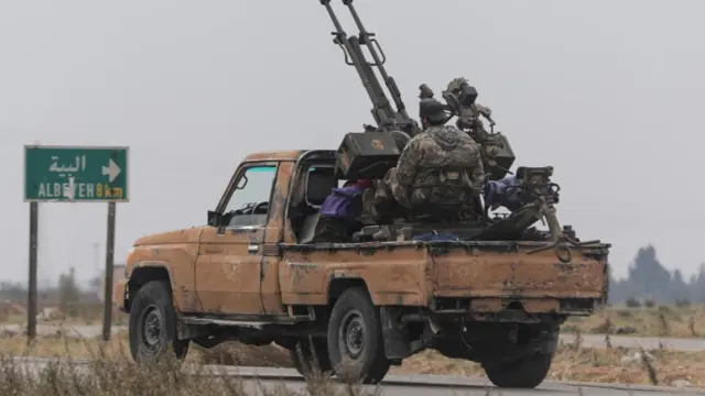 A man sat on the back of a van in the countryside, with a road sign in front, in uniform with a large weapon attached to the van