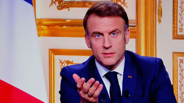 Emmanuel Macron addresses the nation in televised speech after the collapse of the Barnier government. He's sitting down in a mid-shot picture wearing a dark blue suit, matching tie and white shirt. A French flag is visible to his right