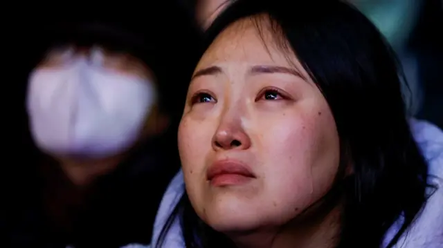 Close up of a woman in light blue jacket as she watches vote results on a big screen during the protests in Seoul. Tear tracks are visible on her face as she looks distraught