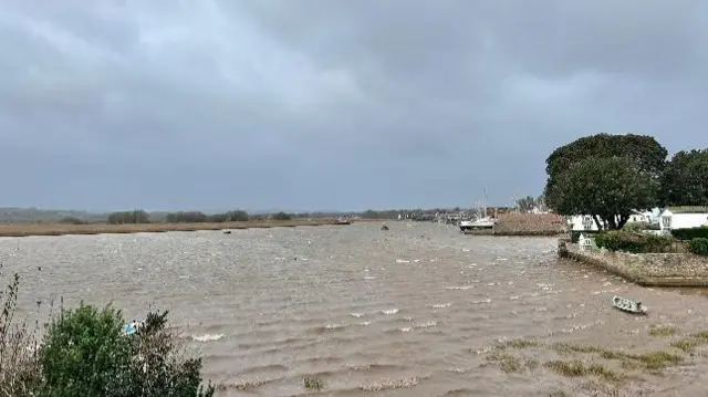 A choppy brown river with several moored boats and trees by the river banks.