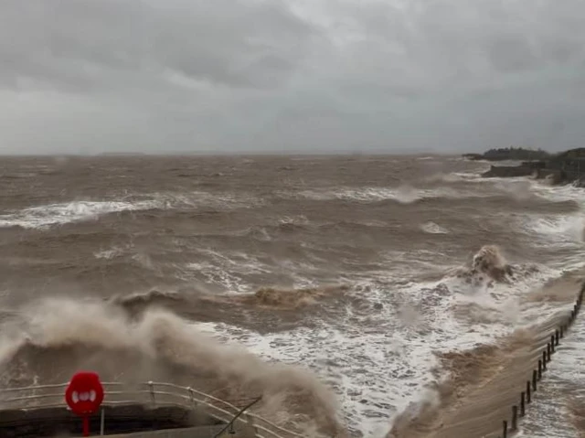 The seafront in Weston-super-Mare. The water is choppy and the waves are high. A red rubber ring can be seen by the railings in the bottom of the frame. The sky is very grey, cloudy and bleak.