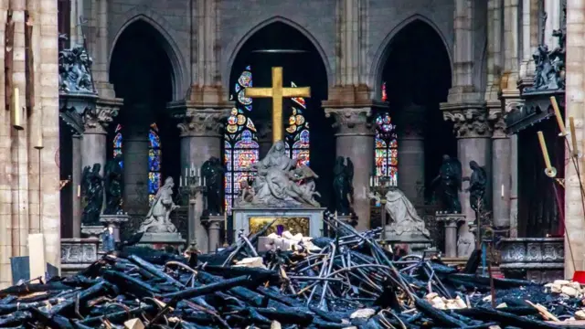 Rubble, debris, and black timber are all piled up on the floor in front of three statues and a golden Christian cross. The statues are grey.