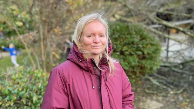 A blonde haired woman wearing a burgundy winter jacket is stood in front of the fallen tree in her garden