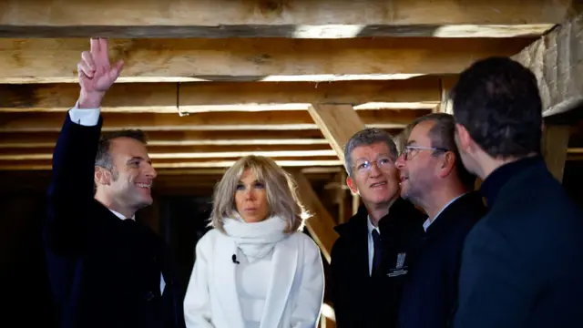 Macron and his wife stand listening to three men. Macron has his hand up touching wood beams above them while smiling. His wife, Brigitte Macron, is wearing a white coat, scarf, and jumper, while the men are all wearing navy coats.