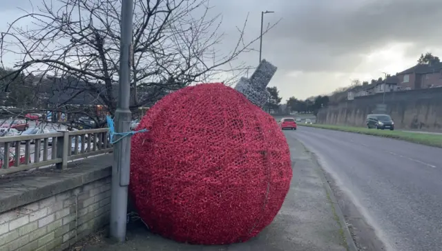 The giant red bauble is tied to a lamp-post on a footpath to the side of the road.