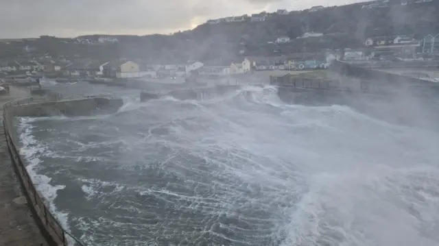 Waves and sea spray tumble in to a harbour wall. The spray has created a mist in the air.