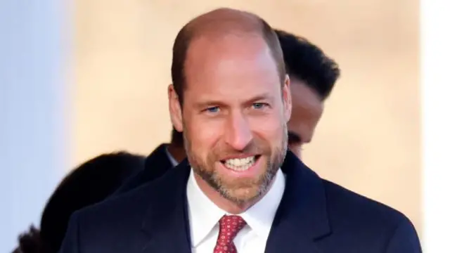 Prince William in dark blue morning coat, white shirt and red tie - mid shot, cuts off below the shoulders. He's smiling as he walks during an official outing in London