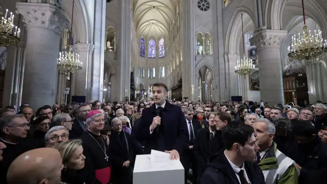 Macron stands in the centre of a crowd in the middle of the Notre-Dame Cathedral under its high ceilings. He is stood at a small podium while holding a microphone. He is wearing a suit and tie with a navy coat.