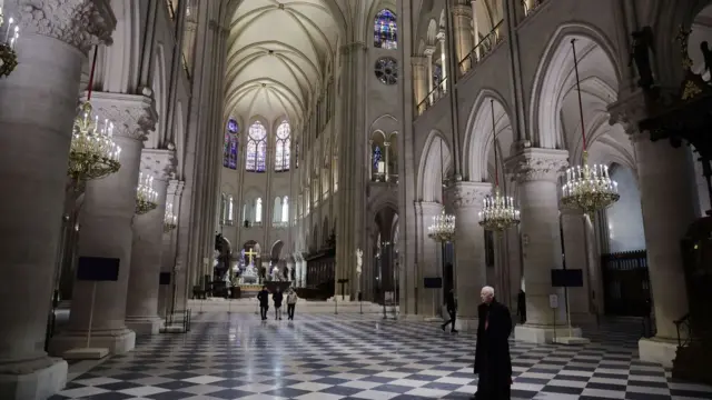 The nave of the cathedral with a few people walking on its black and white squared-floor. There are large arches and a high vaulted ceiling. The stonework is a cream colour and light is coming from several golden chandeliers and purple stained-glass windows