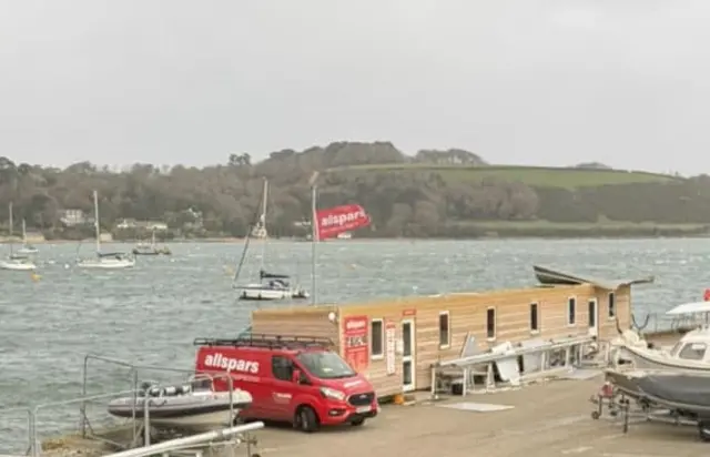 A single-storey wooden building is missing its roof. The sea and opposite mainland are in the background and a parked van and a boat in the foreground.