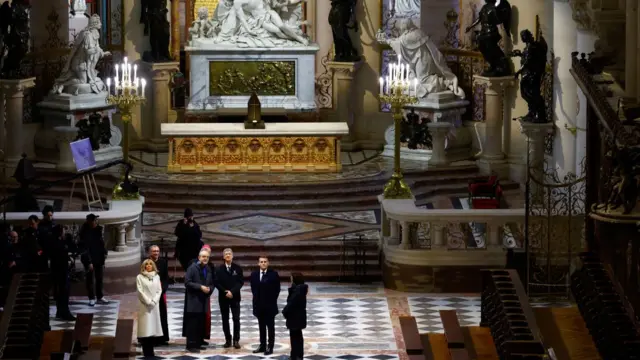 A high up view of the Macron’s tour group looking at statues. The statues are large and white, two depicting men and one depicting a woman. The tour group is stood on a black and white checked floor.