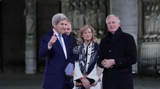 John Kerry holds right thumb up while wearing a blue suit and light blue tie. Isabelle Barnier (C) is in a white coat with a blue patterned scarf around her shoulders while Michel Barnier (R) is in a heavy black coat and scarf