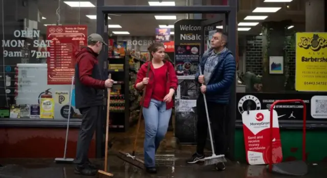 Three people stand in front of a Nisa Local in Pontypridd. They are pushing water with brooms