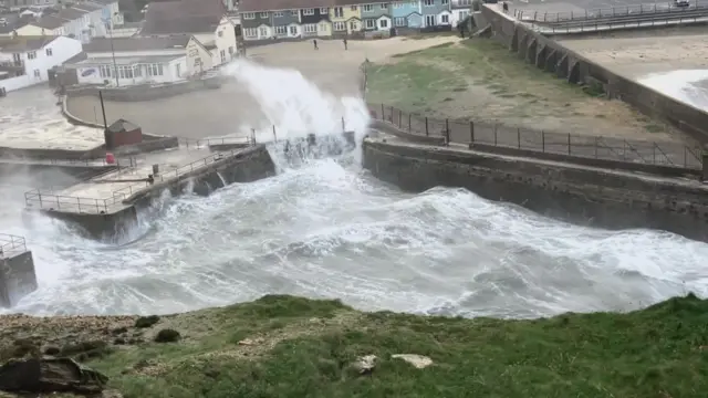 Water spray over a harbour wall with some buildings in the background. The sea is churned up.