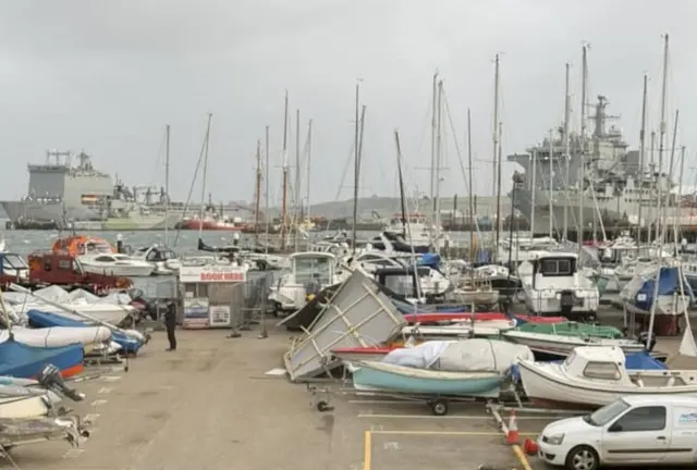 The boatyard is full of laid-up boats. A section of roof has been blown across the yard and is resting on a boat. Royal Navy boats can be seen across the sea in the docks in the background.