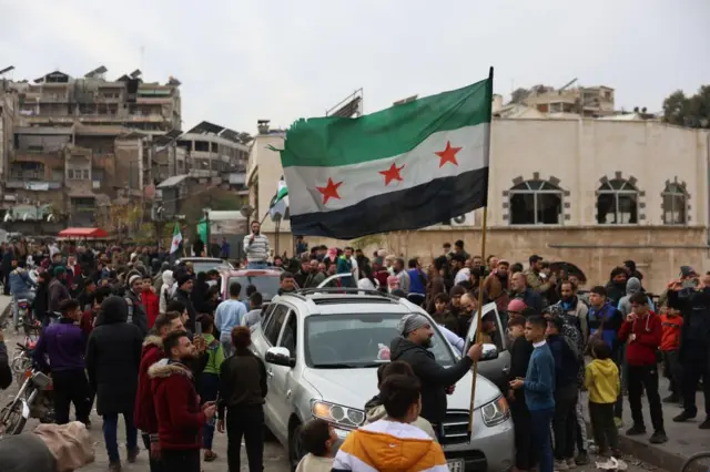 Wide shot showing crowds in Hama around three cars in a line with buildings in the background and a man holding a Syrian rebel flag. Taken 7 Dec