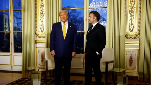 Donald Trump and Emmanuel Macron before bilateral meeting at Elysee Palace. They're both standing in a heavily decorate room, Trump (L) is wearing a dark blue suit and yellow tie while Macron (R) is in a black suti and matching tie with his left hand in his pocket