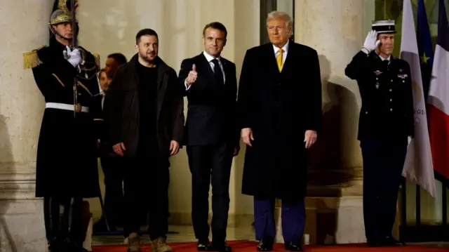 Volodymyr Zelensky, Emmanuel Macron and Donald Trump stand outside Élysée Palace for photos after their meeting. Zelensky is in a black coat, t-shirt and trousers with boots while Macron and Trump are in dark suits