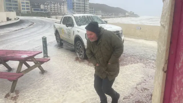 A man in a green jacket battling the wind on a seafront with a truck parked in the background.