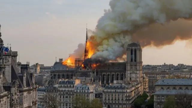 Flames engulf spire of Notre-Dame cathedral after fire erupts inside. Clouds of billowing white smoke can be seen in the sky with the skyline of Paris in the background