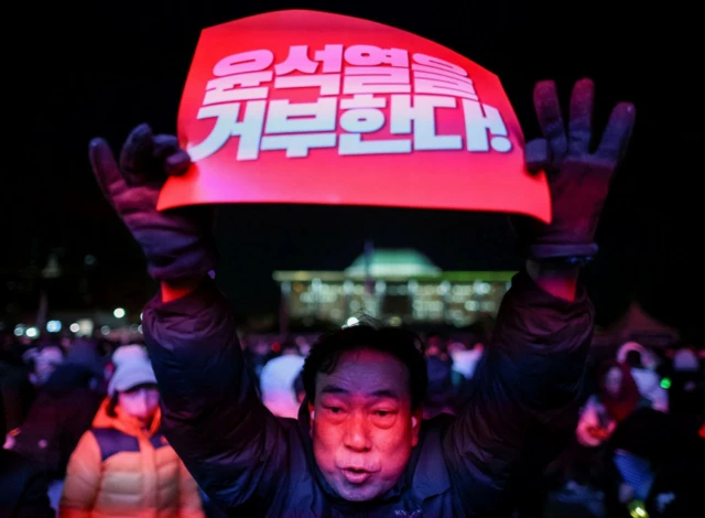 Man holds up a placard demanding the impeachment of South Korean president Yoon. He is wearing gloves and a dark jacket and appears to be mid chant, other protesters (blurred) are visible behind him