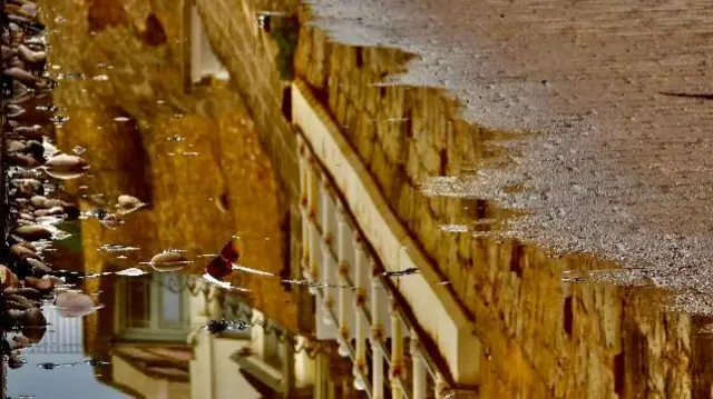 Reflections of houses are seen in a puddle on a brick path with pebbles to one side