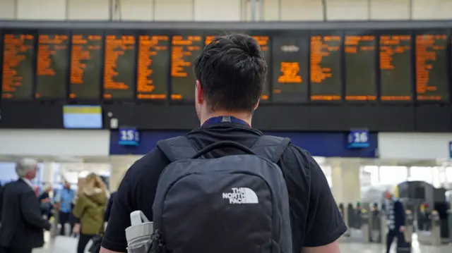 A file photo of a man waiting at a train station with the departure board in the background