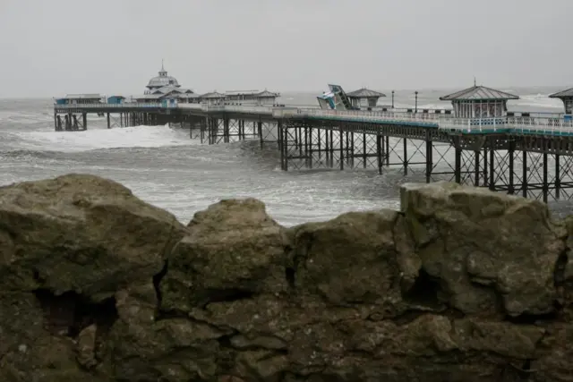 Wider shot of Llandudno pier in North Wales, with rock wall in foreground. One of the huts has been blown over into its side, resting on another hut. Stormy waves and grey skies.