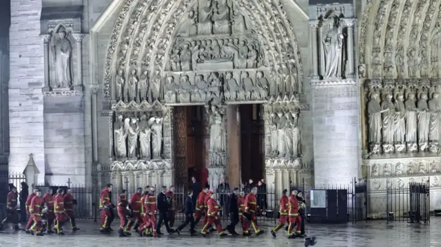 Group of firefighters arrive at Notre-Dame in uniform to attend the reopening ceremony. They're walking in front of one of the side doors of the cathedral, stone decorations and statues illuminated by lights from below