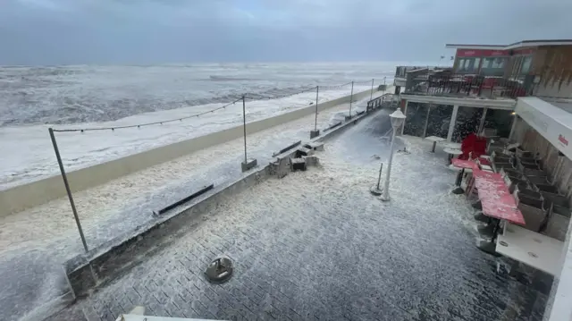 A bird's eye view of a promenade with sea foam on it and a choppy sea in the background.