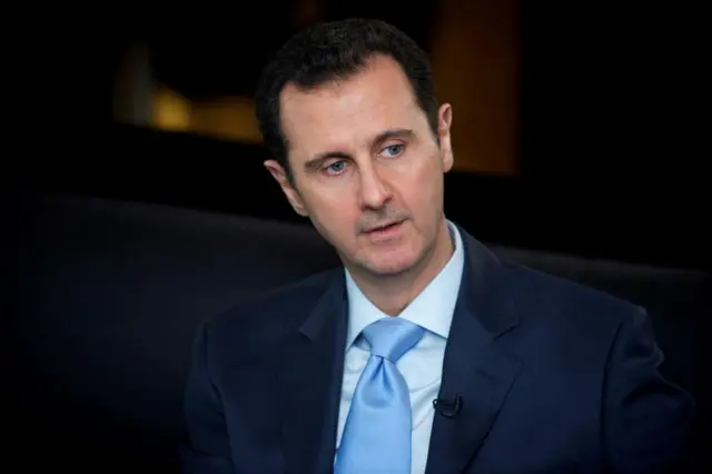Bashar al-Assad in a suit and tie, sat down, looking to the right of the camera with mouth slightly open, dark background