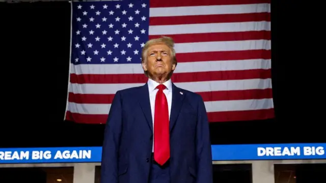 Donald Trump in suit and red tie with large US flag behind him