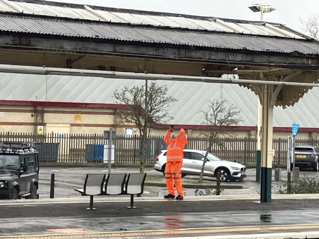 A man in an orange high vis suit standing on the platform at Chippenham station. He is looking up at the canopy with his phone held above his head.