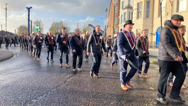 A line of men wearing maroon sashes marching along a road with some buildings in the background.