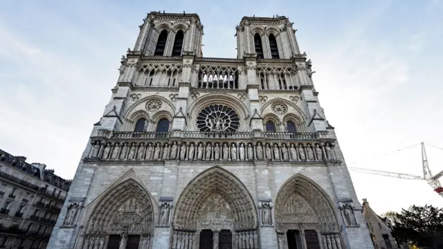 A view of the façade of Notre-Dame Cathedral. It has three arches with six tall doors on the bottom, a row of sculptures above that, and then a third level with two more arches, which contain windows, and a circular window pane in the centre. Above that are two stone towers.