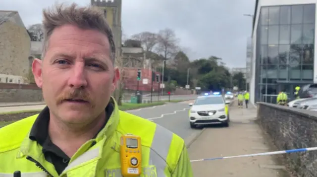 Jonathan Brooker is wearing high-viz uniform and is standing on the footpath along Tregolls Road next to the PKF Francis Clark building. There is a police car and uniformed officers behind him and police incident tape.