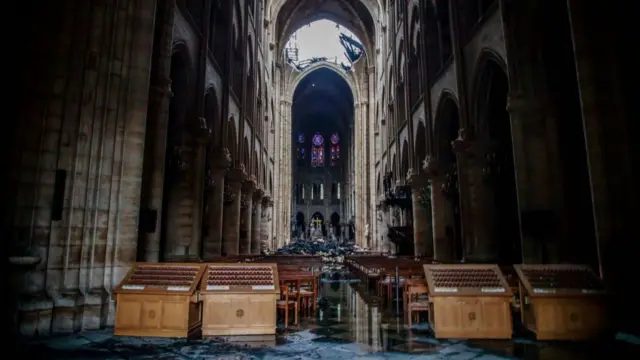 Rubble and debris are on the floor of the cathedral in front of rows of wooden pews, with the sky visible through the large parts of the roof, which is visibly burnt