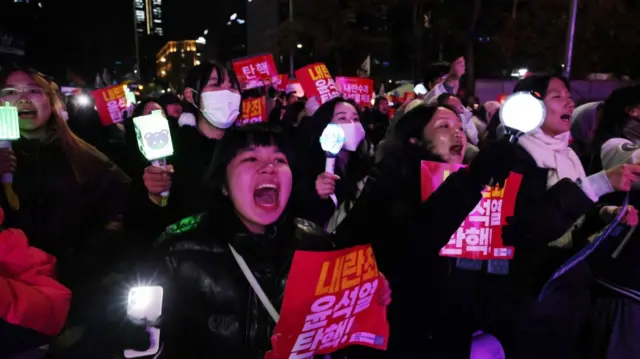 Young women holding placards and candlelights as they demand the impeachment of South Korean president Yoon. A woman at the forefront appears to be shouting and is holding a phone with the flashlight on.