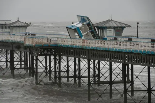 A hut on Llandudno pier in North Wales is blown over and resting on its side against another hut. The pier, with blue and white painted railings, is seen in front of a grey sky and stormy waves.