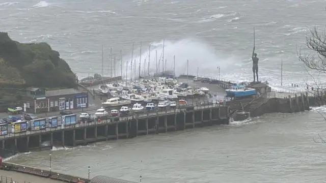 A boat yard with several boats out in a dry dock that is surrounded by water and a hill. A wave is crashing into the dock.