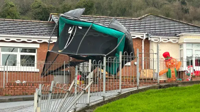 A trampoline upside down and bent out of shape, resting on the garden fence of a house