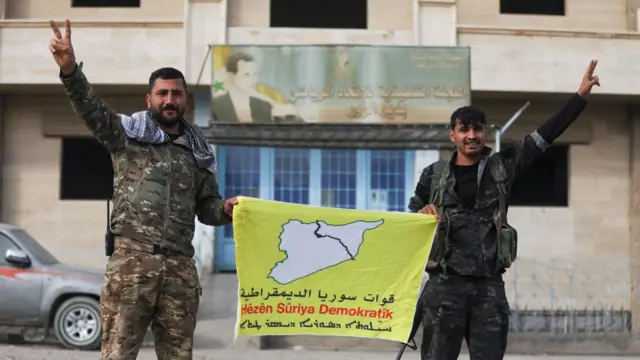 Two men holding up a yellow banner with map of Syria on it, each holding an arm up