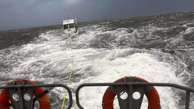 A small pleasure craft tethered to the back of a lifeboat by a tellow rope, There are two red rings on the back of the lifeboat