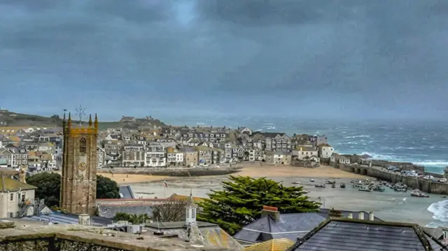 Dark cloudy skies over a beachside town. There is a church belltower in the foreground.